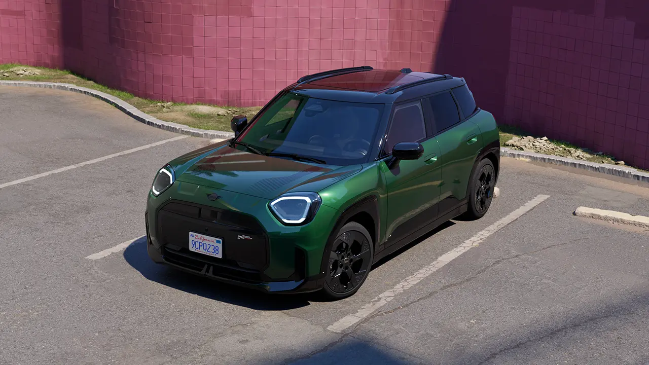 Frontal visning af en parkeret, fuldelektrisk MINI Aceman i British Racing Green på en parkeringsplads med en lyserød væg i baggrunden