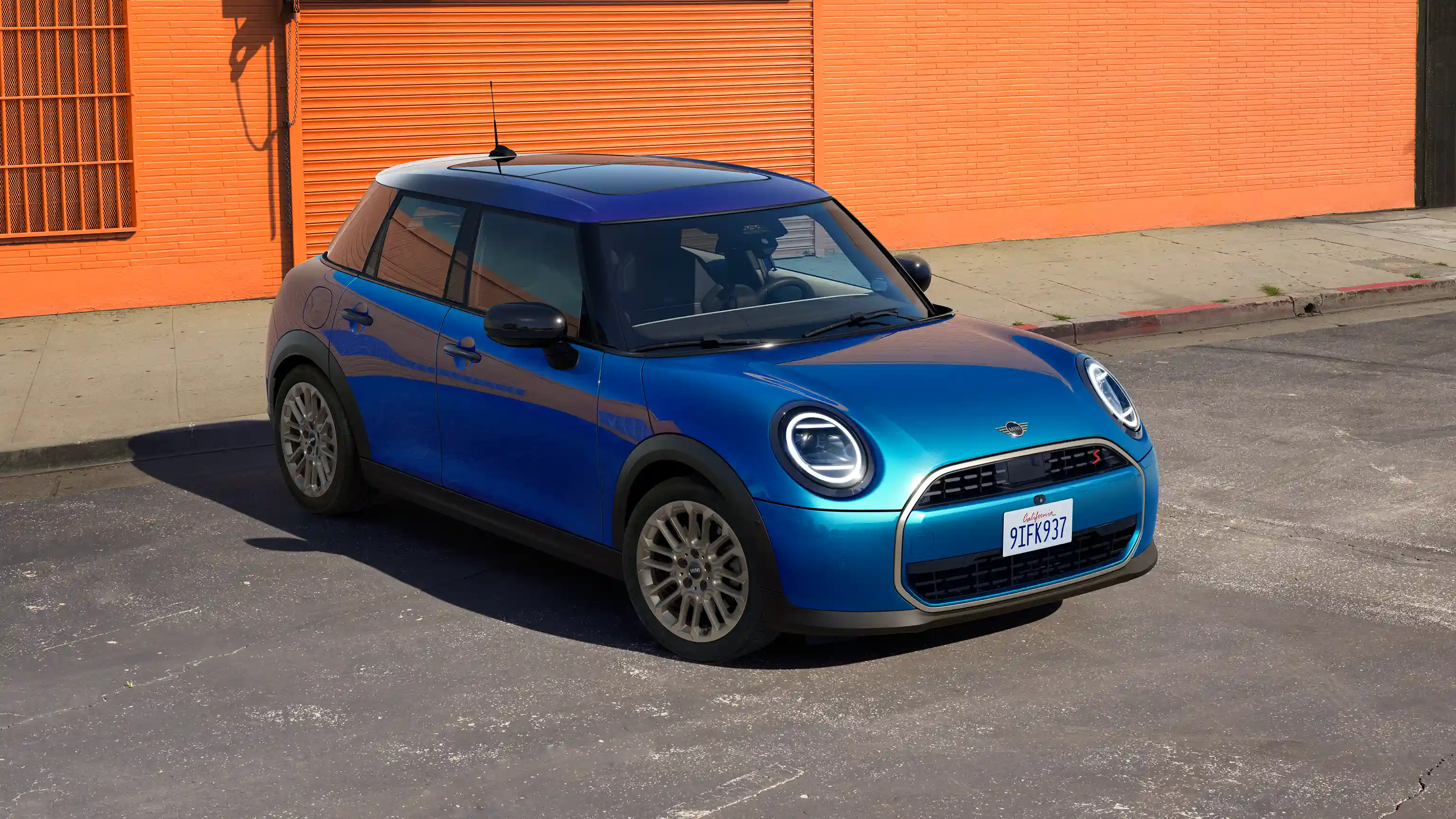 Billede af en parkeret MINI Cooper 5-dørs i Icy Sunshine Blue og tag i Multitone Blue på en gade med en orange væg i baggrunden