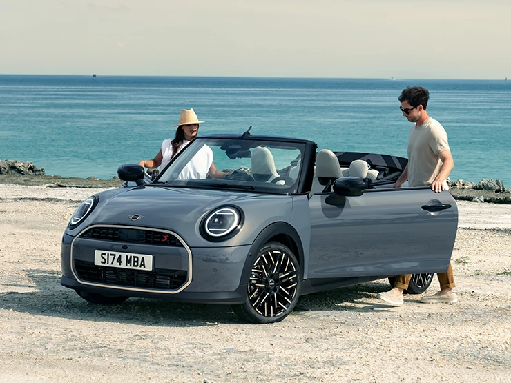Venstre side af MINI Cooper Cabrio set forfra i Copper Grey på en strand med to personer ved siden af.