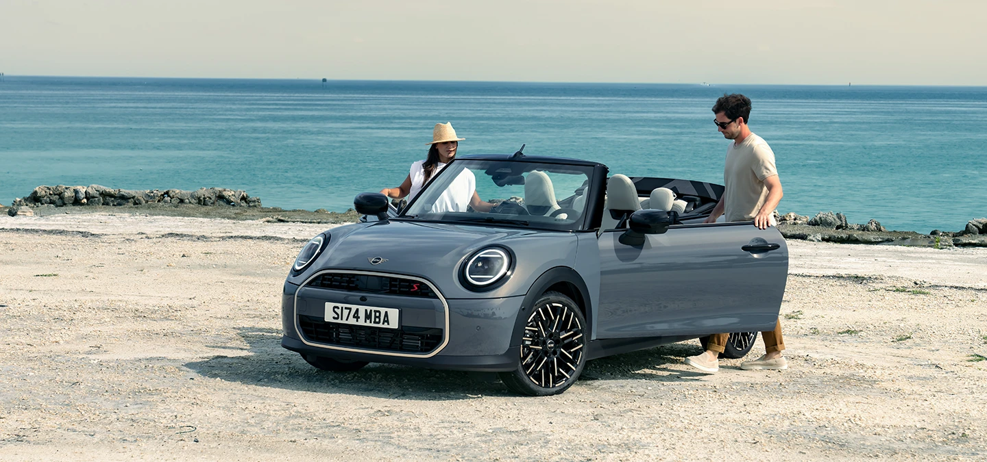Venstre side af MINI Cooper Cabrio set forfra i Copper Grey på en strand med to personer ved siden af.