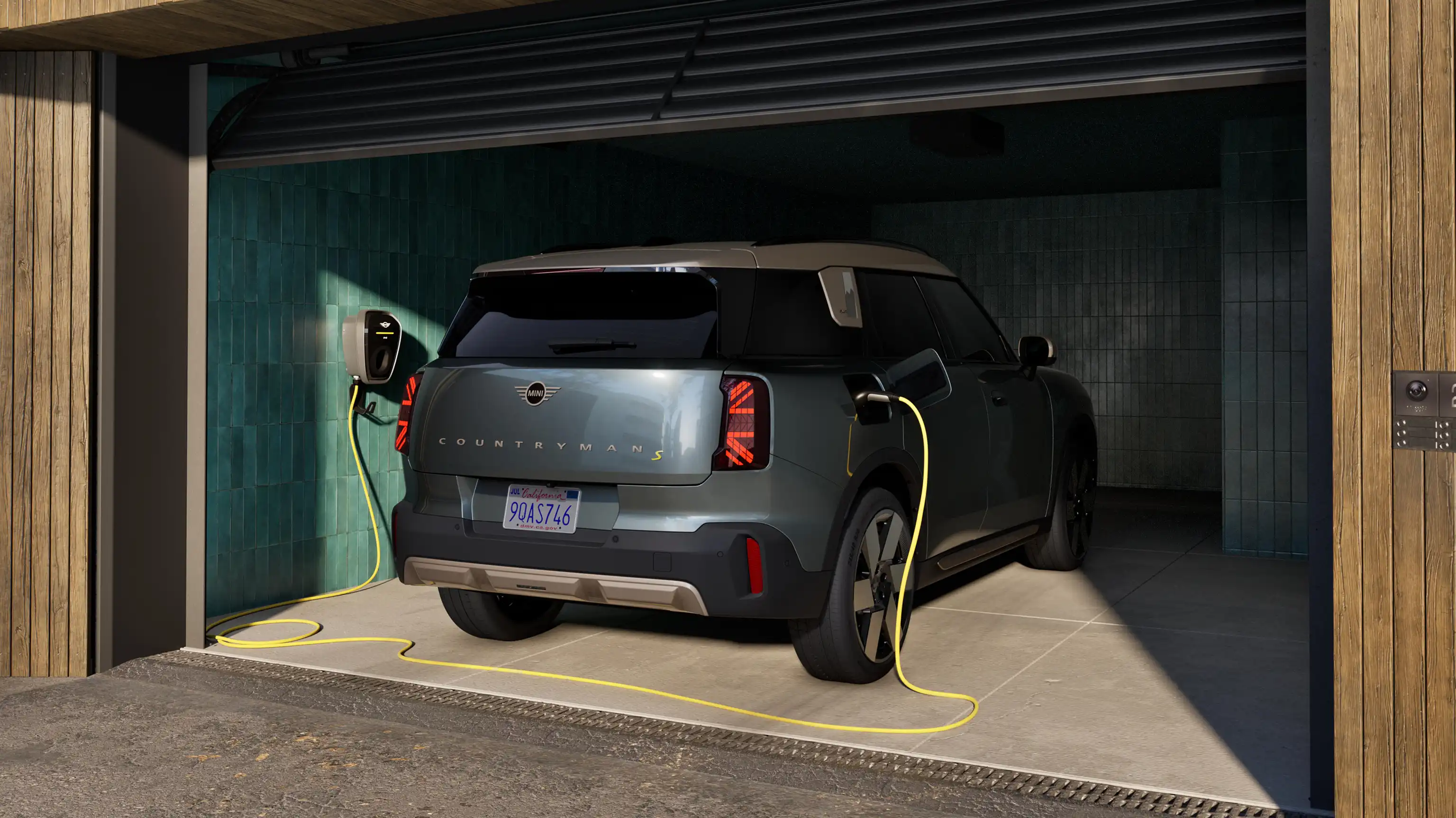 Fuldelektrisk MINI Countryman i Smokey Green, der oplader i en carport og er forbundet med et ladekabel - set bagfra.