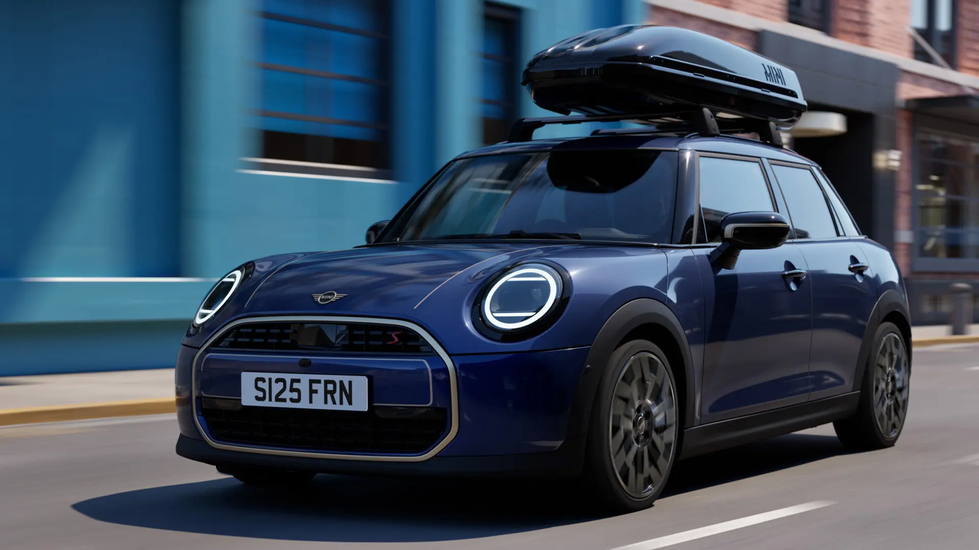 Visning skråt forfra af MINI Aceman i Indigo Sunset Blue med MINI Tagbøjler, der bærer en MINI Tagboks 320. 