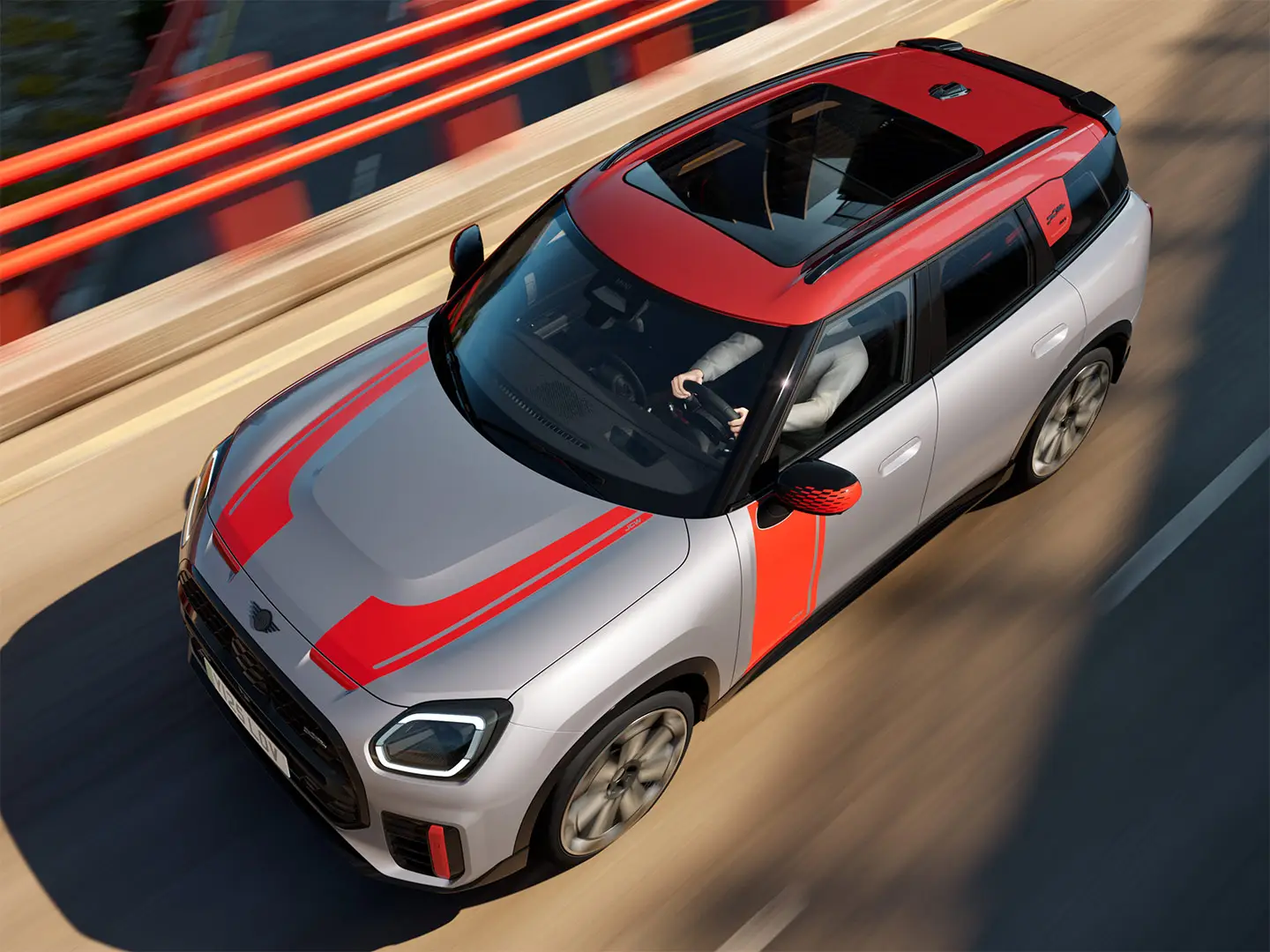 MINI Cooper John Cooper Works i Melting Silver med spejlkapper og fartstriber fra John Cooper Works på motorhjelmen og siden, tagspoiler, og winglet i sort, kørende gennem byens gader – set skråt forfra.
