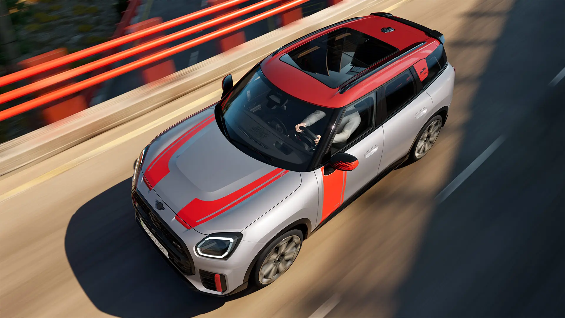 MINI Cooper John Cooper Works i Melting Silver med spejlkapper og fartstriber fra John Cooper Works på motorhjelmen og siden, tagspoiler, og winglet i sort, kørende gennem byens gader – set skråt forfra.
