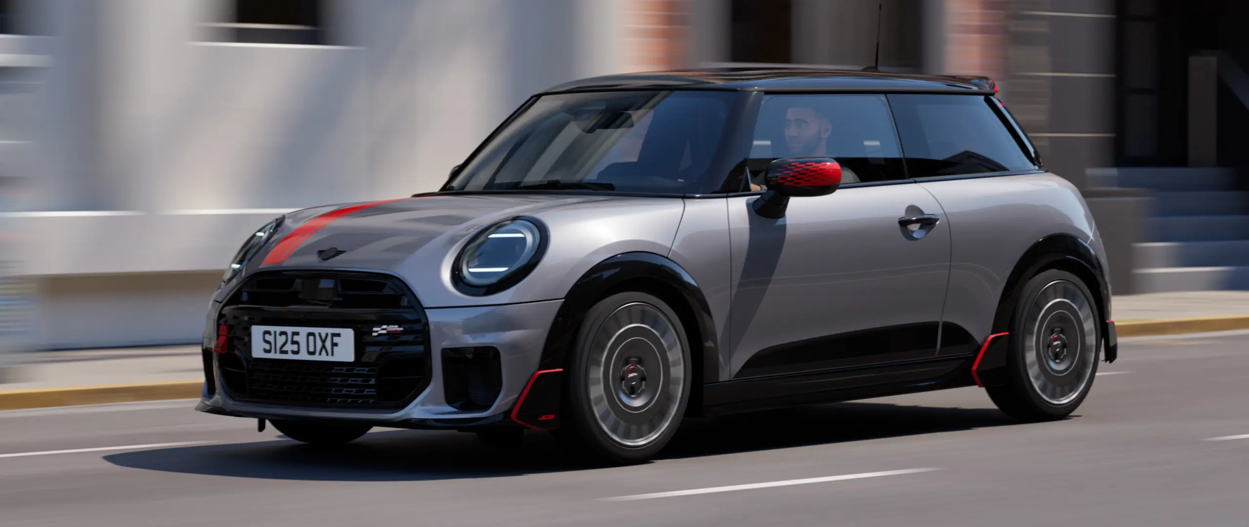MINI Cooper 3-dørs i Melting Silver med John Cooper Works spejlkapper og fartstriber på motorhjelmen i Chili Red, tagspoiler og winglet i sort, kørende gennem byens gader – set skråt forfra.