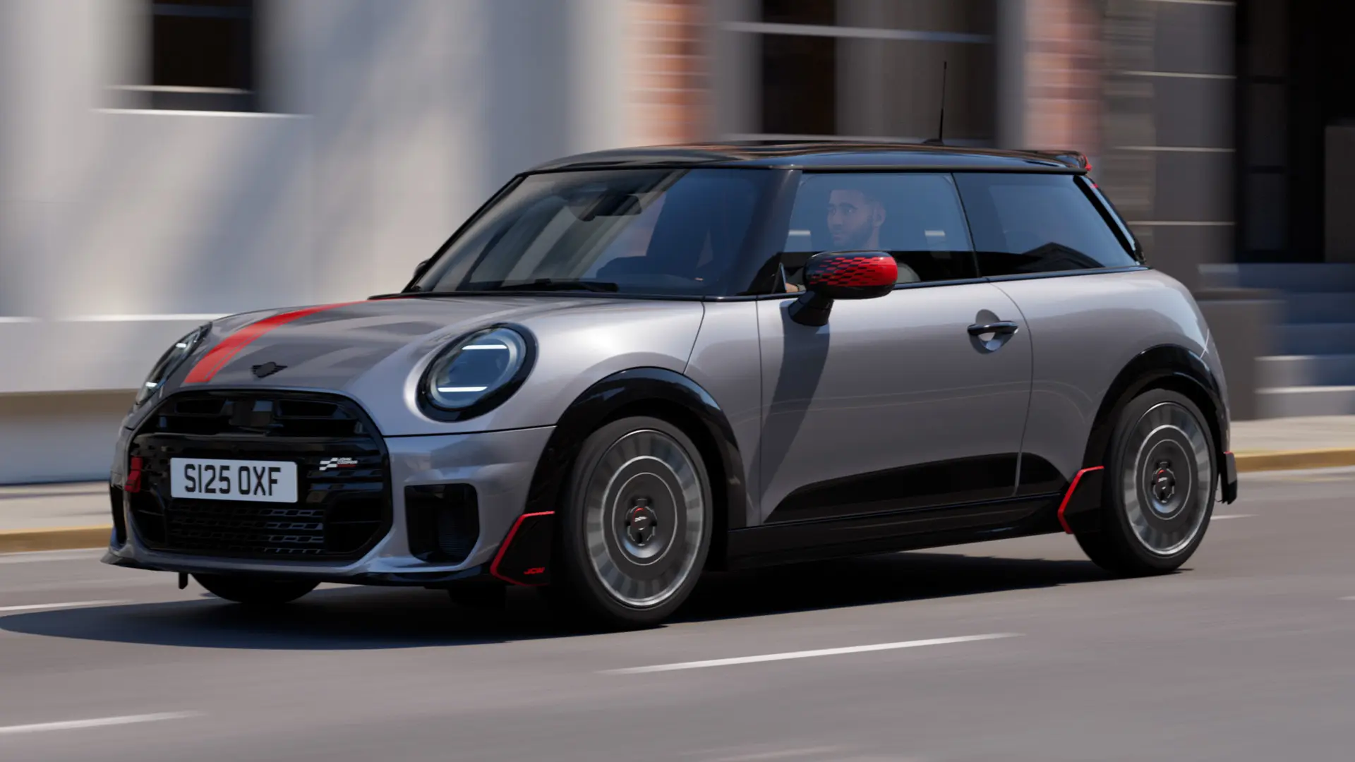 MINI Cooper 3-dørs i Melting Silver med John Cooper Works spejlkapper og fartstriber på motorhjelmen i Chili Red, tagspoiler og winglet i sort, kørende gennem byens gader – set skråt forfra.